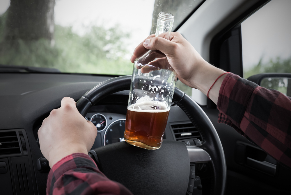 Driver holding a bottle of alcohol while steering a car, symbolizing a drunk driving accident case in Clinton, Massachusetts.