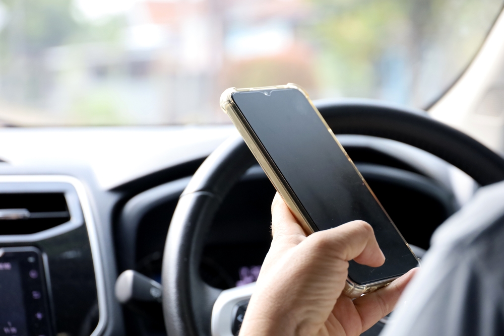 Driver holding a smartphone near the steering wheel, representing distracted driving caused by cell phone use.