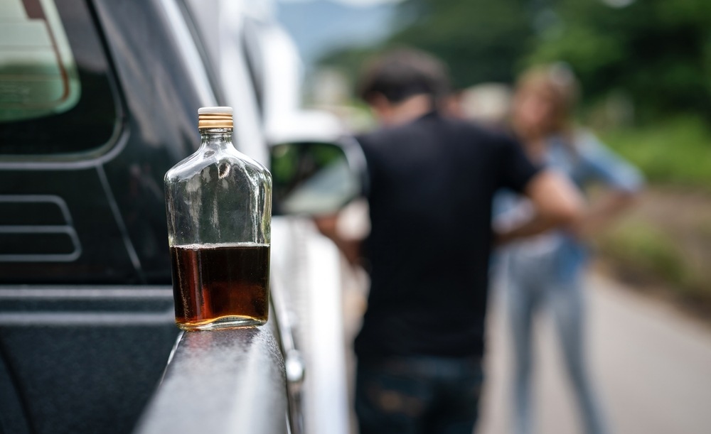 Open liquor bottle resting on the bed rail of a pickup truck while two people talk beside the road in the background